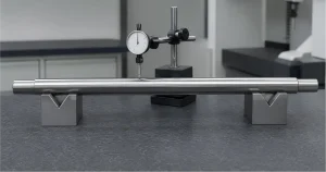 Supported CNC-machined shaft checked for straightness with a dial indicator on a granite inspection table